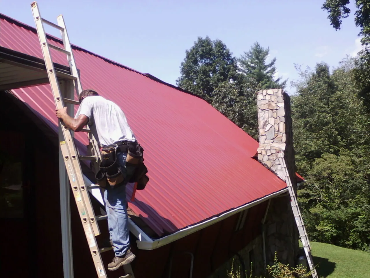 Roofer on a ladder beside a red metal roof and stone chimney — Soffit & Fascia Repair by Roofing Brothers in Aulander